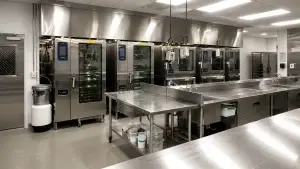 Clean modern stainless steel commercial kitchen with large ovens, prep tables, sinks, hanging equipment, and bright overhead lighting.