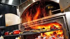 Commercial charbroiler oven with open flames and sparks under a restaurant kitchen exhaust hood during high-heat cooking